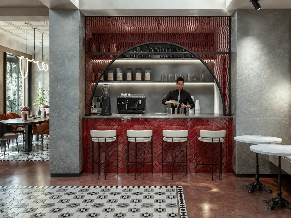 Kahve barı karşıdan: kemer biçimli kırmızı metal kafes niş, altında espresso makinesi ve çalışan barista; önde balık sırtı kırmızı seramik bar ve tabureler